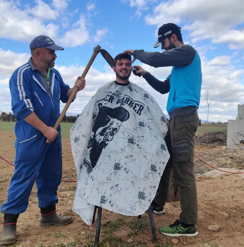 Escena de un corte de pelo al aire libre con tres personas y el campo de fondo
